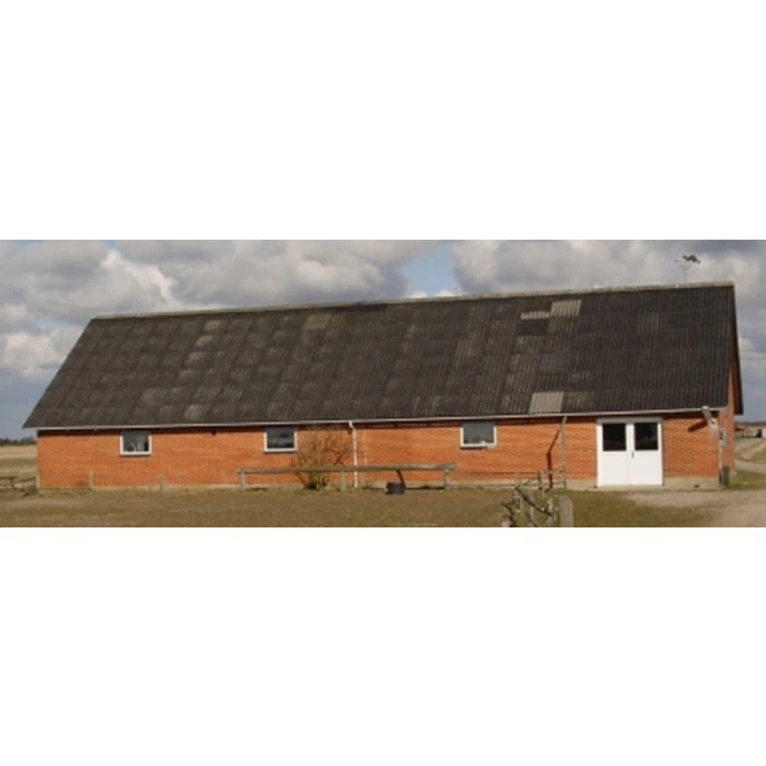 En lang, enetages rød murstensbygning med et stort, mørkt skråt tag, der ligger på åben mark under en delvist overskyet himmel.